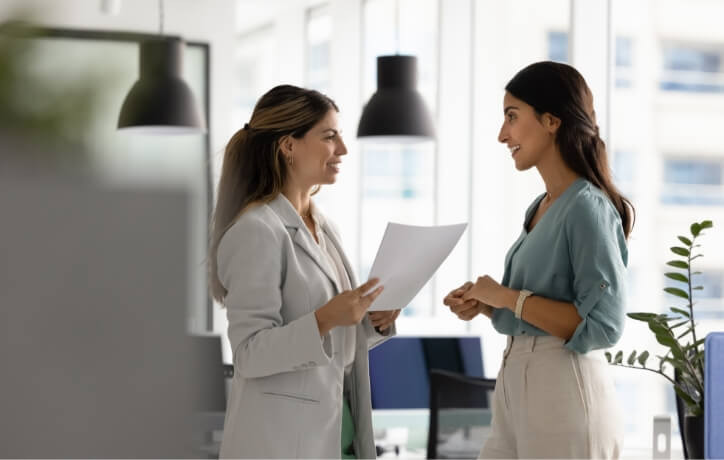 Two women professionals discussing business documents in a modern office with city views and hanging lamps