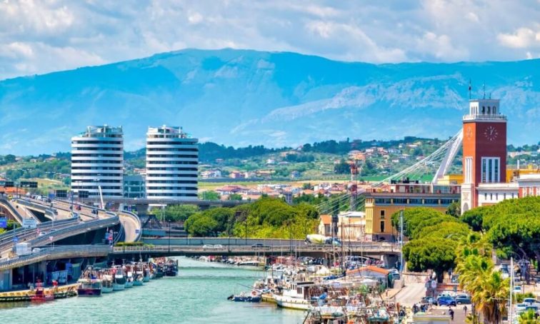 Coastal city with modern high-rise buildings, harbor, bridge, and mountains under blue sky