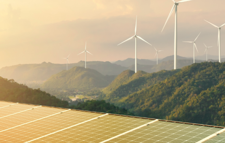 Solar panels in foreground with wind turbines on forested hills at sunset, symbolizing renewable energy solutions