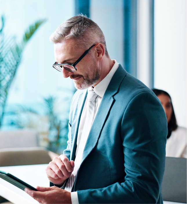 Professional man in teal blazer reviewing tablet in modern office with plant and colleague visible